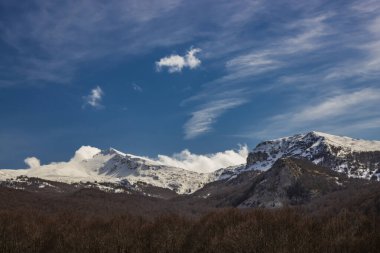 İtalya, Campo Felice 'deki Abruzzo Apennines' in karla kaplı dağları. Aşağıda, kışın çıplak ağaçlarla dolu bir kayın ormanı var. Kayalık dağların tepelerindeki beyaz bulutlar.