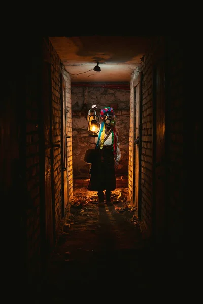 戴着防毒面具的乌克兰女人拿着一盏旧煤气灯点亮了路 穿着传统的民族服装 他们躲藏在地下室的炸弹掩蔽所里 躲避化学攻击 毒气和辐射 — 图库照片