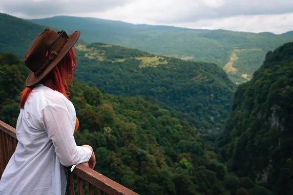 stock image Woman traveler in Okatse Canyon in Georgia, standing on hanging metal pedestrian pathway trail above deep precipice. Leather hat. Travel and vacation. Back view. copy space