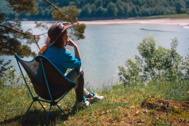 Kız göle bakan dağdaki manzaranın tadını çıkarıyor. Portatif kamp sandalyelerinde oturuyorlar. Turizm ve tatil için ekipman kavramı. Etkin yaşam biçimi