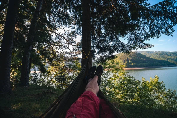 Man in hammock, first person look view, warm summer day, pine tree. river and mountains background. Travel and vacation, tourism equipment concept. Copy space