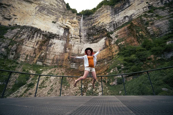 Girl jumping in happiness on mountain waterfall at summer cloudy day time enjoy view with kinchkha waterfall, Georgia. Concept of tourism and travel.