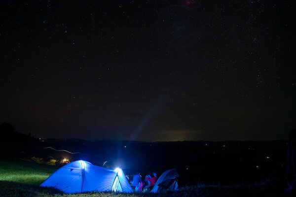 Голубая палатка с горящим светом стоит на поляне под звездным небом. На горизонте горы Альп. Концепция звездного ночного неба и Млечного Пути. кемпинг в горах.