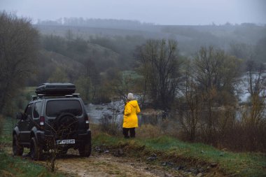 Khmelnytskyi, Ukrayna - 15 Nisan 2023: Suzuki Jimny off-road aracı, koyu gri, çatı rafı Niken 390, ilkbahar günü, nehir manzarası. Ekstrem maceralar için bir araba. Sarı ceketli kadınlar.