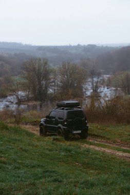 Khmelnytskyi, Ukrayna - 15 Nisan 2023: Suzuki Jimny off-road aracı, koyu gri, çatı rafı Niken 390, serin bahar günü, nehir manzarası. Turizm ve aşırı maceralar için bir araba. Dikey fotoğraf
