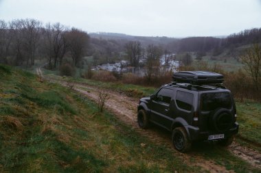 Khmelnytskyi, Ukrayna - 15 Nisan 2023: tepedeki Suzuki Jimny off-road aracı, koyu gri, çatı rafı Niken 390, serin bahar günü, nehir manzarası. Turizm ve aşırı maceralar için bir araba.