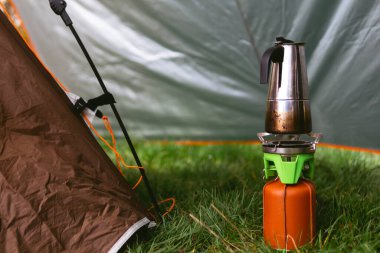 Küçük bir gaz ocağı, kahverengi deri kapaklı bir yakıt silindiri, gayzer kahve makinesi. Sahada yemek pişirmek. Taşınabilir kamp malzemeleri, turistik malzemeler. Bahar günü. Yeşil çimenler. Çadır. Seçici odak