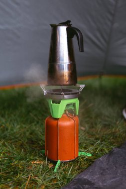 Modern bir gaz ocağı, deri kapaklı bir yakıt silindiri, gayzer kahve makinesi. Sahada yemek pişirmek. Taşınabilir kamp malzemeleri, turistik malzemeler. Bahar günü. Yeşil çimenler. Dikey fotoğraf.
