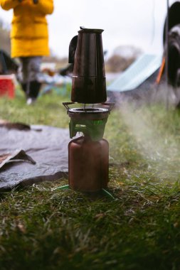 Seçici odaklanma. Gaz ocağı, deri kapaklı bir yakıt silindiri ve gayzer kahve makinesi. Sahada yemek pişirmek. Taşınabilir kamp malzemeleri, turistik malzemeler. Bahar günü. Yeşil çimenler. Dikey fotoğraf.