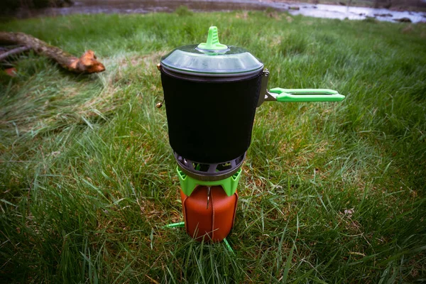 stock image Close up. A modern gas burner, a fuel cylinder, a kettle with a protective cover. Cooking in the field. Portable camping, tourist equipment. Spring day. Green grass.