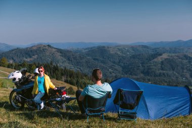 Bir motorcu, bir erkek ve bir kadın. Derin bir bakış açısıyla dinlen. Sevgi dolu iki turist. İlişki ve macera kavramı. Motosiklet, çadır. Aktif yaşam tarzı. Katlanan sandalyeler. Üst dağlar.