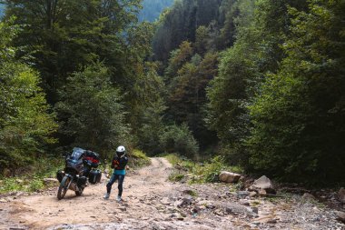 Özel motosikletli kız, koruma, dizlik ve balıkçı yaka. Beyaz kask. Şehirlerarası bir motorsikletin yanında duruyor. Yan çantalar ve bavullar. Dağ yolu. Boşluğu kopyala.
