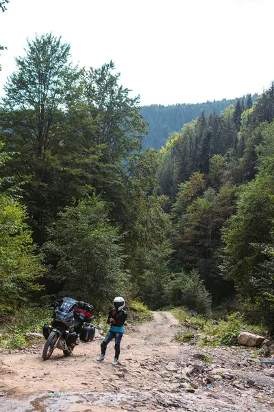 Özel motosikletli kız, koruma, dizlik ve balıkçı yaka. Beyaz kask. Şehirlerarası bir motorsikletin yanında duruyor. Yan çantalar ve bavullar. Dağ yolu. Dikey fotoğraf.