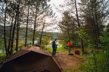 Bir erkek ve bir kadın bir turist çadırının yanına katlanan sandalyeler kurdular. Kozalaklı orman, arka planda arazi aracı, dinlenme ve tatil, Dinyester nehir kıyısı, Ukrayna.