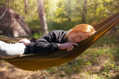 Çam ormanının ortasında hamakta dinlenen bir kadın gün batımını izliyor. Yavaş yaşam konsepti. Hippi. Kamp çadırı. Yakın plan. Kameraya bak.