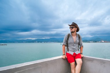 Kırmızı şortlu, sırt çantalı, şapkalı ve güneş gözlüklü bir adam rıhtımda oturuyor ve manzaranın tadını çıkarıyor. Karadeniz Batumi, Gürcistan