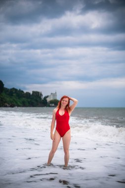Kırmızı kapalı mayolu genç bir kadın. Büyük dalgalı fırtınalı deniz, ufukta şiddetli fırtınalar. Gün batımı. Rocky plajı. Dikey fotoğraf.