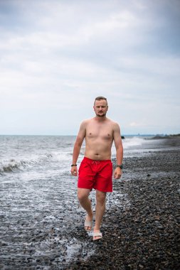 Kızıl şortlu bir adam Karadeniz 'in kayalık sahillerinde yürüyor. Büyük fırtına dalgaları ve yağmur bulutları. Tatilciler için tehlike. Vahşi Ortanca plajı. Dikey fotoğraf