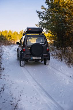 Khmelnytskyi, Ukrayna - 30 Kasım 2023: Suzuki Jimny off-road aracı, dört çeker. Çatı rafı. Ormanın ortasında, karlı bir yolda duruyor. Arka plan Yedek tekerlek korunuyor. Dikey fotoğraf