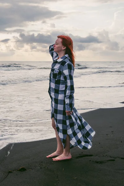 A young woman in a long checkered shirt stands on the black sand near the water edge, stormy waves of the sea. Heavy gray rain clouds at sunset. Vertical photo.