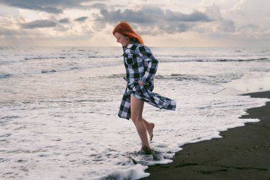 Uzun damalı gömlekli genç bir kadın siyah kumun üzerinde duruyor. Soğuk suda ayaklar, denizin fırtınalı dalgaları. Gün batımında yoğun gri yağmur bulutları.