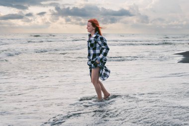 Uzun damalı gömlekli genç bir kadın siyah kumun üzerinde duruyor. Soğuk suda ayaklar, denizin fırtınalı dalgaları. Gün batımında şiddetli gri yağmur bulutları. Ureki Sahili, Georgia