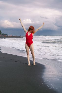 Kırmızı elbiseli mutlu kadın turist. Siyah kumların üzerinde, denizdeki büyük fırtınalı dalgalar. Yüzmek için tehlike. Ufukta yağmur bulutları var. Ureki Sahili. Georgia mı? Dikey fotoğraf