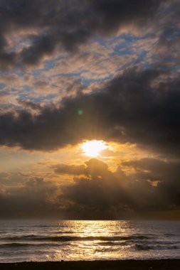 Gün batımı manzarası, deniz kıyısı, fırtınalı dalgalar, ufukta sağanak yağmur bulutları, güneş bulutların arasından bir göz gibi kırılıyor. Dikey fotoğraf