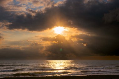 Gün batımı manzarası, deniz kıyısı, fırtınalı dalgalar, ufukta sağanak yağmur bulutları, güneş bulutların arasından bir göz gibi kırılıyor.