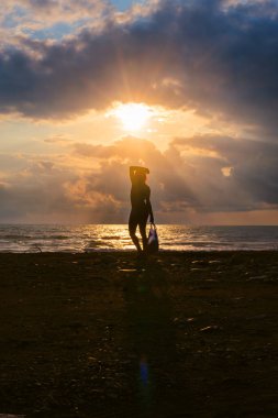 Gün batımında bir paket taşıyan bir kadının silueti, deniz kıyısı, arka planda fırtınalı dalgalar, ufukta sağanak yağmur bulutları, güneş bulutların arasından bir göz gibi çıkıyor. Dikey fotoğraf.
