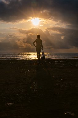 Gün batımında bir paket taşıyan bir kadının silueti, deniz kıyısı, arka planda fırtınalı dalgalar, ufukta sağanak yağmur bulutları, güneş bulutların arasından bir göz gibi çıkıyor. Dikey fotoğraf.