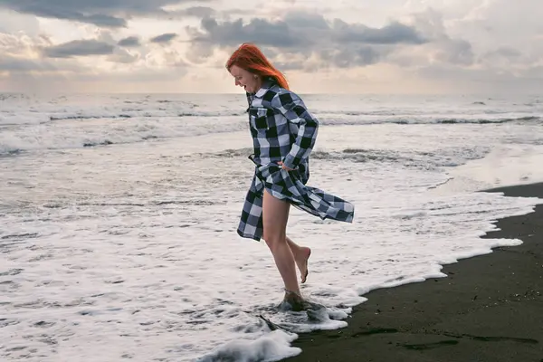 A young woman in a long checkered shirt stands on the black sand. feet in cold water, stormy waves of the sea. Heavy gray rain clouds at sunset.