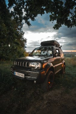 Khmelnytskyi, Ukrayna - Ağustos. On bir. 2024: Eski Jb43 Suzuki Jimny 2010 açık hava. Çatı rafı. Bir off-road aracı izleme noktasında park halinde. Yaz günü, ağaçların arasında gölgeler. Dikey fotoğraf
