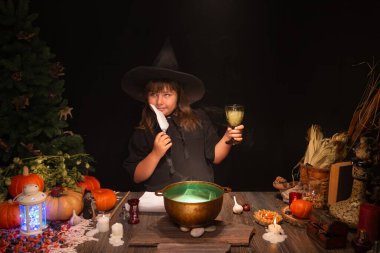 Halloween, smiling little cute girl dressed as witch in witch's hat brews a glowing magic potion in cauldron on coals. Magic wand, andles, pumpkins, ingridients, fog smoke, magic, mystical atmosphere clipart