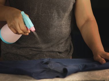 A person's hand holds a pink and blue spray bottle, releasing a mist onto a dark blue textile.