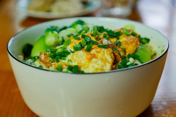 Close-up of the food inside the white porcelain bowl. A white porcelain bowl on a wooden table with a lot of food inside, with slightly browned poached eggs at the top, and some green vegetables and scallions. It tastes delicious and looks good.