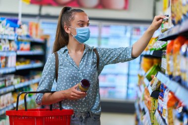 Tıbbi koruyucu maskeli bir kadın marketteki bir raftan yiyecek ürünlerini seçiyor ve alıyor. 