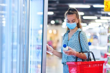 Tıbbi koruyucu maskeli, alışveriş sepetiyle sıradan bir genç bayan alıcı marketteki dondurucudan süt ürünleri seçiyor. 