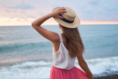 Beyaz bluzlu, pembe etekli ve hasır şapkalı, romantik bir kadın deniz kıyısında dikiliyor ve uzaklara bakıyor.       