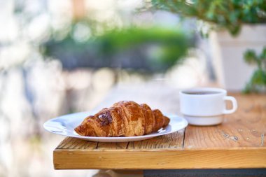 Coffee cup and fresh baked croissant on a plate on a wooden table for french breakfast in the morning on a balcony