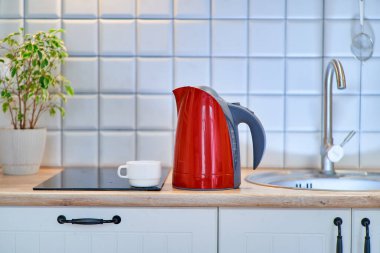 Evdeki mutfaktaki masada beyaz bardaklı modern kırmızı elektrikli su ısıtıcısı.    