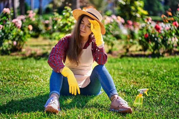 暖かい晴れた日に家の庭で草の上に休んで暑い天候に苦しんでいるわらの帽子 エプロンと黄色のゴム手袋の疲れ女性の庭師 — ストック写真