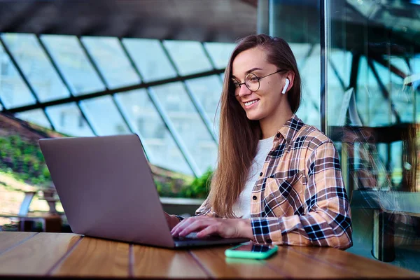 ワイヤレスヘッドフォンやメガネの幸せな女性のフリーランサーは カフェでオンラインでリモートで作業するためのラップトップを使用しています デジタルスマートモビリティライフスタイルを持つ現代人 — ストック写真