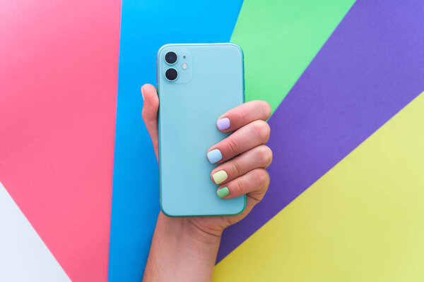 Female hands with color manicure holds green mint colored phone with dual lenses on a bright colorful background. Top view 