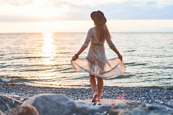 stock image Boho chic woman in long fluttering dress and felt hat standing back on stone by the sea at sunset                