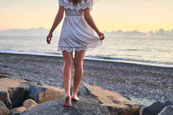 Romantic woman with long legs in a short summer white dress standing on a stone by seashore at summertime