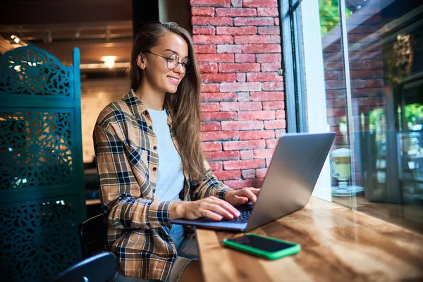 年轻而聪明的现代女性自由职业者 戴着眼镜 在咖啡店的笔记本电脑上远程工作 舒适的工作场所 — 图库照片