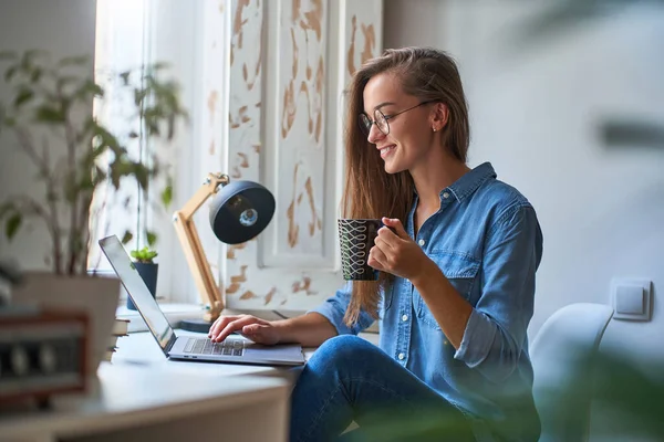 快乐而随意的年轻女性 戴着圆圆的眼镜 带着一杯咖啡在电脑前在线浏览 舒适舒适舒适的工作环境在窗边 — 图库照片