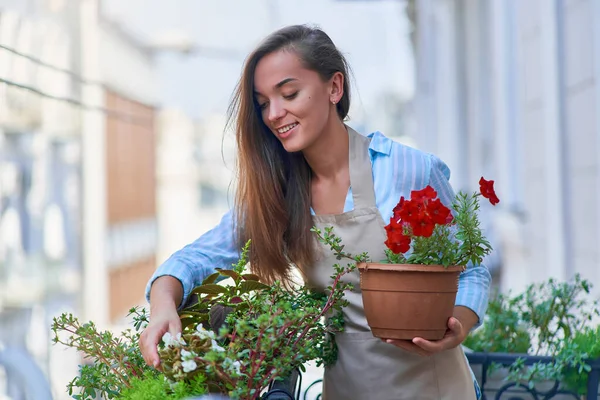 幸せな笑顔かわいい女性庭師エプロン保持フラワーポットペチュニアを身に着けているとバルコニー植物について世話をする — ストック写真
