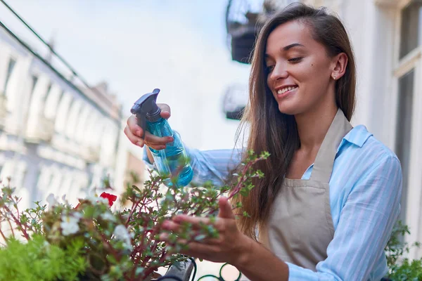 スプレーボトルを使用してエプロン散水バルコニーの花を身に着けている幸せな笑顔かわいい女性庭師 — ストック写真